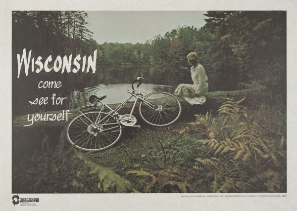 Wisconsin Division of Tourism poster. View toward a woman sitting on a log at the shoreline of a lake or river with a bicycle laying on its side in the foreground. Trees on the far shoreline and plants in the foreground are starting to turn fall colors.   