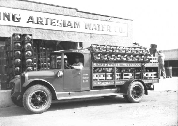 A man is sitting in the driver's seat of a beverage delivery truck. In the background on the right is another man who is standing on a loading dock just behind the truck on the left. The sign on the side of the truck bed reads: "Sparkletts Artesian Water Co." The truck bed is full of crates of large bottles of water. The building behind the truck has a sign painted above the loading dock door that reads, in part: "_ling Artesian Water Co."