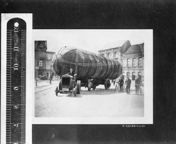A man is driving a tractor to pull a large tank along a street. Two men are standing nearby holding bicycles.