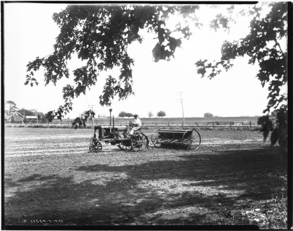 View of a man using a Farmall tractor to seed a field. There are farm buildings in the background.