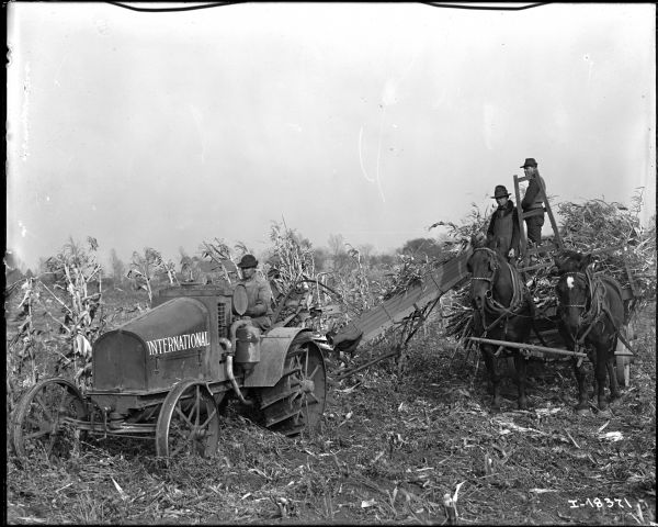 View from front of a man driving an International tractor pulling a corn picker. On the right two men stand on a horse-drawn wagon piled with cornstalks.