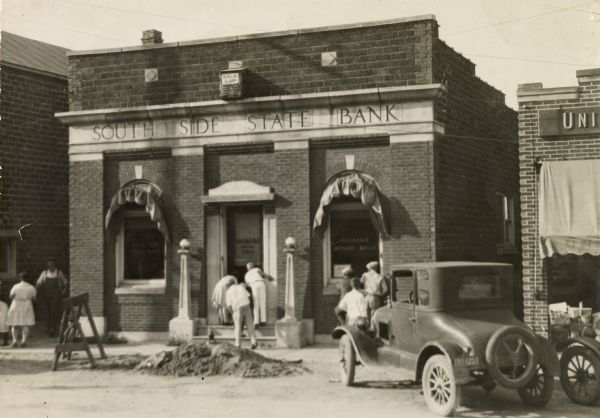 View from street of the South Side State Bank on Lakeside Street, after a robbery. The front door is set in the center of the facade, flanked on both sides by windows with awnings. The name of the bank is above the door, and above that is a box reading: "Burgler Alarm." Three people are standing on the front steps and peering in through the front door. Other people are standing on the sidewalk in front. There is a traffic barrier and a mound of dirt in the street in the street in front of the bank. On the right is the Universal Grocery Co.