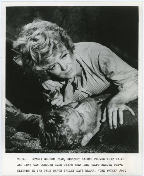 Mary Parker, played by Dorothy Malone, leans over Rafe Pegarski (Steve Clinton) in a scene from the TV show <i>Death Valley Days</i>.  Both are on the ground and dirty.  Malone holds an open pocket watch in one hand.