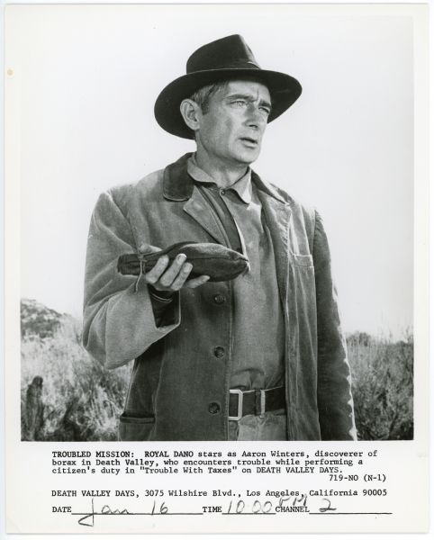 Aaron Winters (Royal Dano) holds a bag in one hand in a promotional still for the episode "Trouble With Taxes" of the TV show <i>Death Valley Days</i>.  He is standing outside and is wearing a hat and coat.