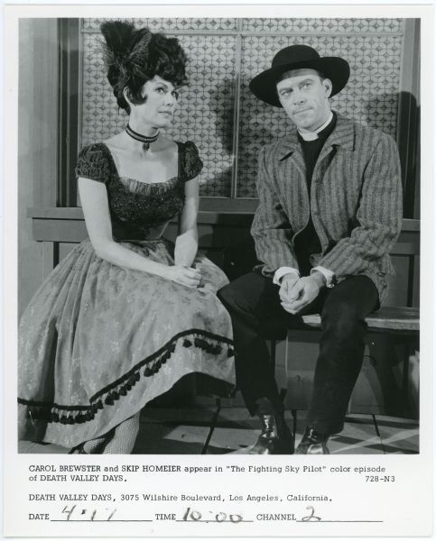 Claire Vernon (Carol Brewster) sits on a wooden bench outside a building next to Reverend Ben Darniell (Skip Homeier) in a scene from the episode "The Fighting Sky Pilot" from the TV show <i>Death Valley Days</i>.  Brewster is dressed as a saloon girl while Homeier wears a black hat and a clerical collar.