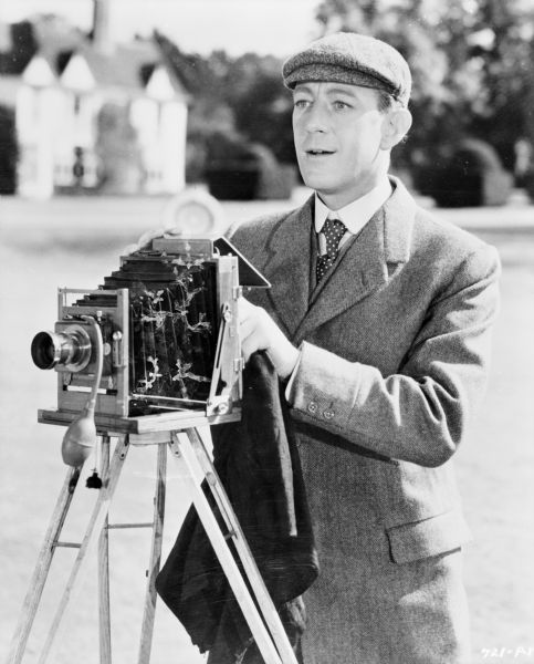 Image of actor Alec Guinness as Young Henry in the 1949 film <i>Kind Hearts and Coronets</i>.  Guinness stands behind a camera as in the film, Young Henry is an amateur photographer.