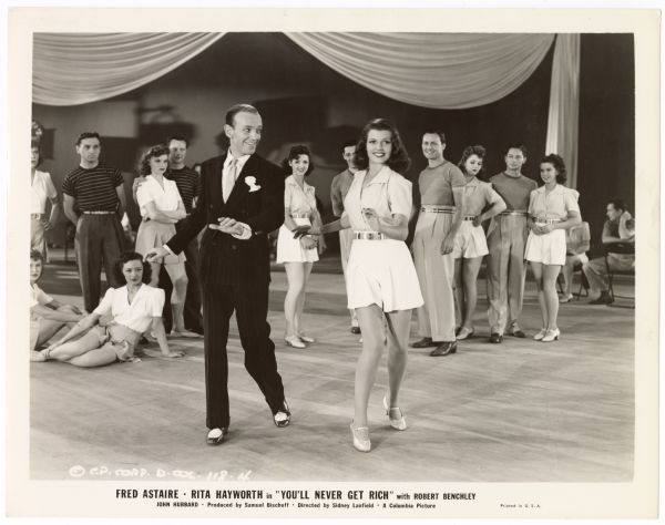 Fred Astaire looks at Rita Hayworth as the two dance in a scene from the 1941 film <i>You'll Never Get Rich</i>.  A group of dancers stand behind them and watch.