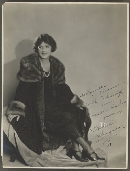 Actress Helen Ferguson sits and poses in a dark fur trimmed coat.  She is also wearing a dark dress with lace at the bottom, dark shoes, and a string of pearls.  The photograph has an inscription from Ferguson to columnist Louella Parsons.