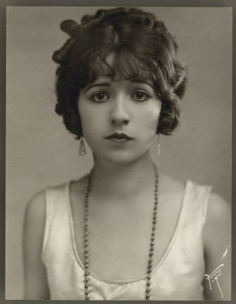 Portrait of a young Helen Ferguson.  She looks directly at the camera and wears a sleeveless light colored dress along with earrings and a beaded necklace.