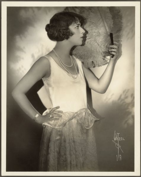 Full length photograph of actress Helen Ferguson.  She stands in profile to the camera with one hand on her hip and the other holding a feather fan.  Ferguson wears a light colored sleeveless dress with a lace skirt along with a double strand necklace of pearls.
