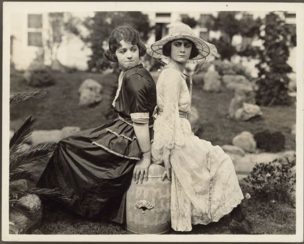 Actresses Helen Ferguson (in dark dress) and Ruth Stonehouse (wearing hat) sit back to back on a milk can.  Both have annoyed/determined looks on their faces.  The pair wear dresses and are outside.