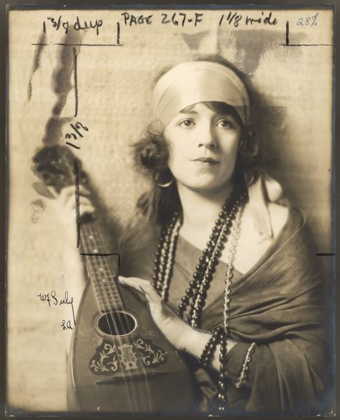 Actress Helen Ferguson holds a mandolin in a publicity photograph.  She wears a headband, hoop earrings, and strands of long beads around her neck.