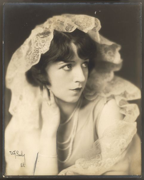 Actress Helen Ferguson poses for a portrait.  She looks off to the side and has one hand slightly touching her chin near her ear.  She has a long lace veil on top of her head which cascades down her shoulder.  She is also wearing four strands of a pearl necklace.