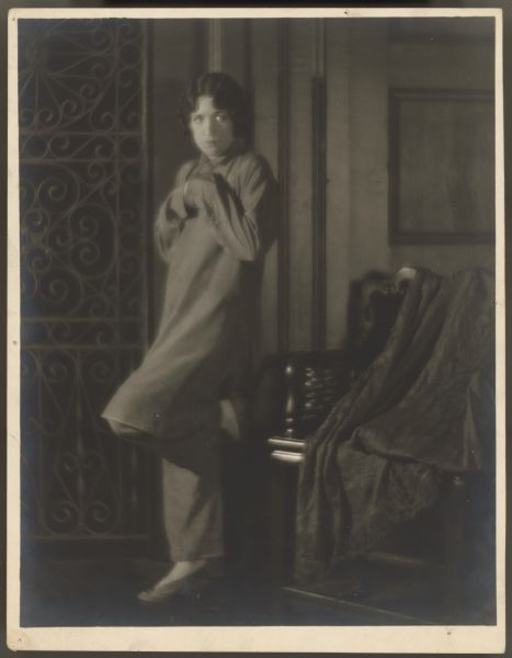 Publicity photograph for the 1923 film <i>The Unknown Purple</i> of actress Helen Ferguson.  She stands with one foot against a wall and wears a long tunic with pants underneath it.  She holds her hands together and hides them in the sleeves of the tunic.