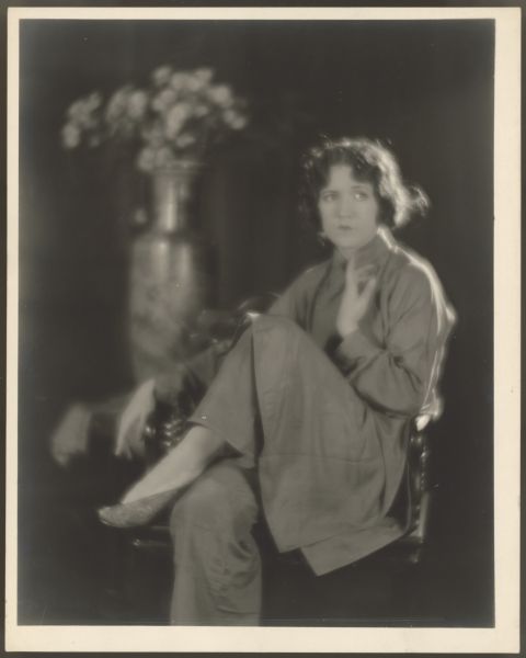 Actress Helen Ferguson sits in a large chair with one leg crossed.  One hand is curled up against her chest while the other rests on the arm of the chair.  She wears a large tunic with pants underneath it.  A large vase with flowers is in the background.