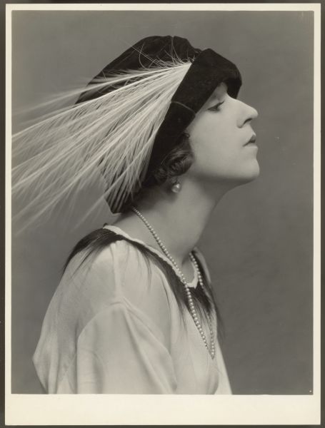 Actress Helen Ferguson sits for a photograph in profile.  She wears a dark colored hat with a large white feather.  Her blouse is white with dark trim (possibly fur).  One earring is visible and she wears a single strand of pearls around her neck.  Her face is tilted upward.