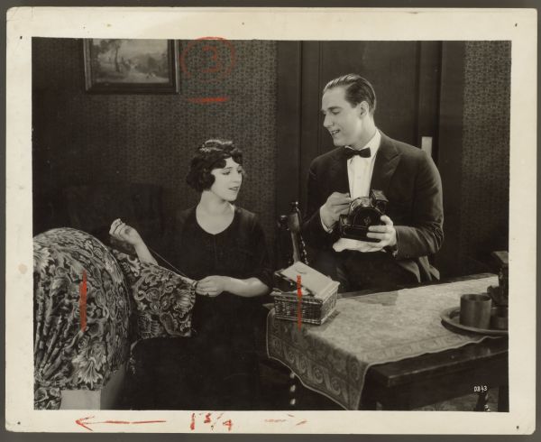 Actress Helen Ferguson and actor David Butler appear in a still from the 1921 film <i>Making the Grade</i>.  Ferguson sits next to a table and sews/mends a large piece of fabric.  Butler sits next to her and holds a toy cash register.