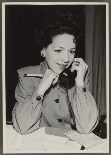 Photograph of actress, now press agent, Helen Ferguson.  She sits at a desk and has a telephone receiver to one ear.  She holds a pencil in the other hand.  Papers are on the desk in front of her.