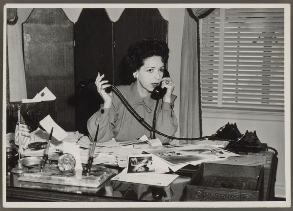 Actress, now press agent, Helen Ferguson sits at a desk and talks on the telephone.  She is holding receivers for two telephones - she holds one to her ear and the other off to the side.  Her desk is full of papers and photographs.  There is a large pen set and a small U.S. flag at the front of the desk.