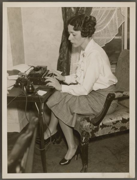 Actress, now press agent, Helen Ferguson sits at a small folding table and types.  A small telephone is next to the typewriter.  Ferguson sits on a nice chair with her legs crossed as she works.  She is wearing a white top with ruffles and a skirt.