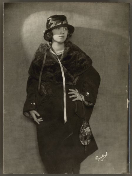 Actress Helen Ferguson poses against a wall.  She wears a dark knee length coat with a large fur lapel.  Two strands of a ribbon lay over the front of the coat.  She wears a brimmed hat a a large decoration on the front and holds a small string bag with one hand. 
