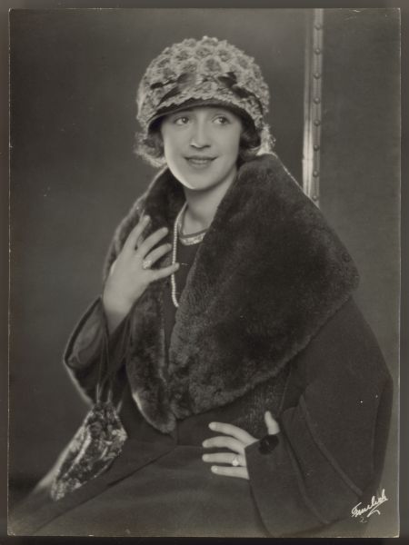 Actress Helen Ferguson sits while wearing a dark coat with a large fur trimmed lapel.  Her hat is covered with small flowers and a ribbon.  A small handbag hangs by strings from one arm which is holding onto the lapel of the coat.  The other hand is on her hip.