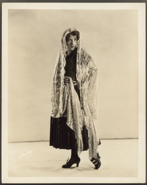 Full length photograph of actress Helen Ferguson.  She wears a dark colored dress with a large scarf or shawl draped over her head and shoulders.  There are large buckles on her shoes.