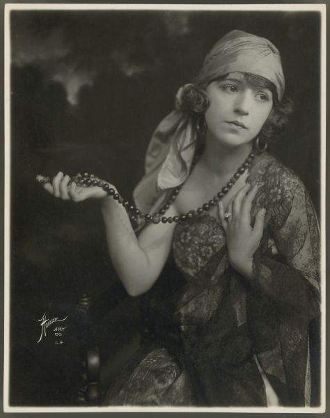 Actress Helen Ferguson poses while holding a large beaded necklace with one hand.  She has a large scarf tied around her head and wears hoop earrings.  Her dress is strapless and is made of a dark lace and she has a dark lace veil/shawl draped over one shoulder and arm.