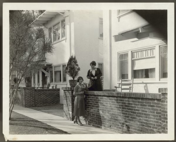 Actress Helen Ferguson stands on the sidewalk next to a brick wall in front of her home in Hollywood.  Her mother stands on the patio on the other side of the brick wall.  Ferguson is holding a small bouquet of flowers.