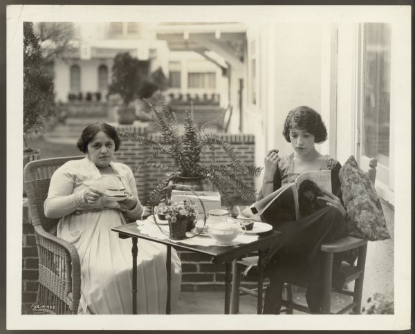 Actress Helen Ferguson and her mother sit on the porch of their Hollywood home.  The two are having something to eat and drink.  Helen reads an issue of <i>Cosmopolitan</i> magazine while her mother looks directly at the camera.  A potted plant is behind them.