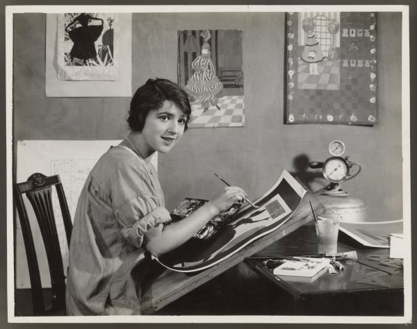 Actress Helen Ferguson sits at a table and works on a poster.  She wears a smock and a small string of pearls.  There are several posters or painting on the wall behind her and paint supplies on the table.