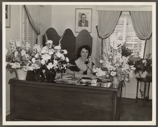 Actress, now press agent, Helen Ferguson sits at her desk and smiles while looking at the camera.  She holds a telephone receiver to one ear and holds a pencil in the other hand.  Three large flower arrangements sit on her desk with more flowers on a table and window sill behind the desk.