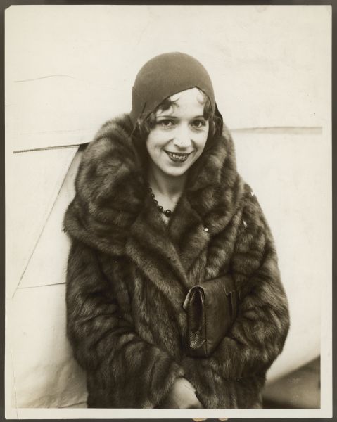 Actress Helen Ferguson poses for a photographer outside while wearing a fur coat.  Her hat covers her head completely with just a little of her hair showing.  She has a handbag tucked underneath one arm.