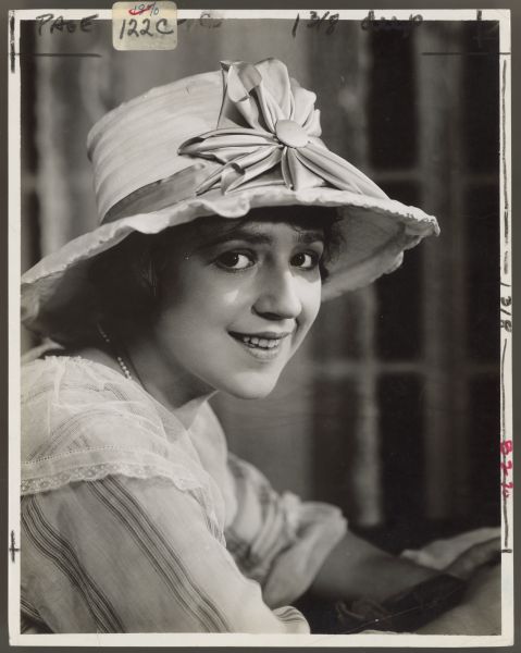 Actress Helen Ferguson looks at and smiles for the camera.  She has a light colored wide brimmed hat with a large bow on the front on her head.  Her blouse or dress has a lace lapel.