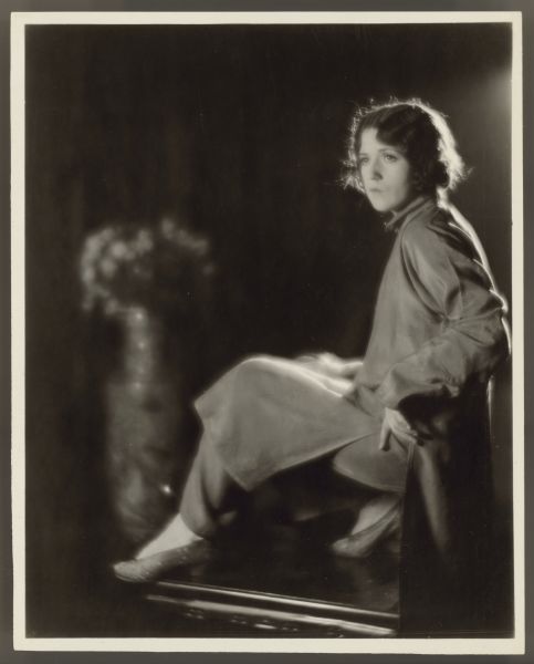 Actress Helen Ferguson sits with one leg stretched out and has a stern look on her face.  She wears a long tunic with pants underneath it.  A blurry large vase of flowers can be seen in the background.