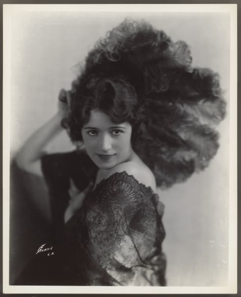 Actress Helen Ferguson holds a large feather fan behind her head while looking directly at the camera.  She wears a strapless dress with dark lace over one shoulder.