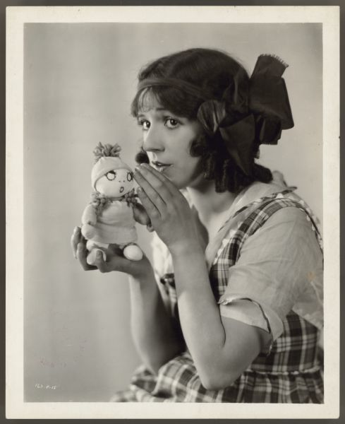 Actress Helen Ferguson holds a small doll in one hand and pretends to whisper something to it.  She is dressed like a young girl in a blouse and jumper and has a big bow in her hair.