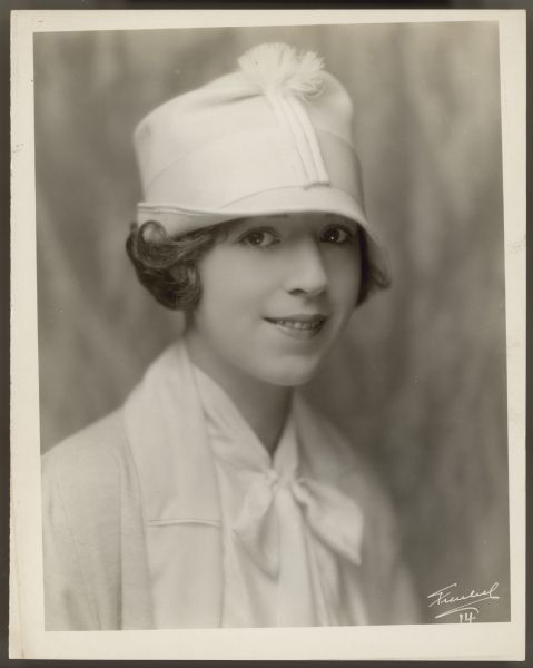 Publicity photograph of Helen Ferguson.  She smiles and looks at the camera while wearing a white hat with a small decoration at the front.  She also wears a white coat and blouse with a bow.