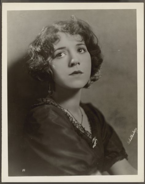 Portrait photograph of actress Helen Ferguson.  She sits and looks up and away from the camera.  Her dress is dark in color with a small amount of lace trim along the V-neck.  A cameo brooch is at the bottom of the V-nect and she also wears a small necklace - possibly with a crucifix.