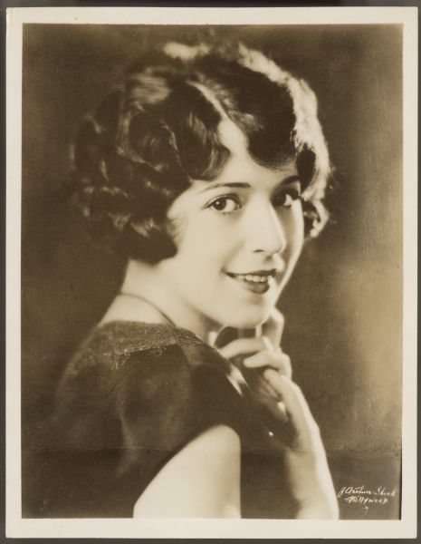 Sepia toned photographer of actress Helen Ferguson.  She looks over her shoulder and smiles at the camera.  Her hands are up by her chin and she is wearing a sleeveless dress.