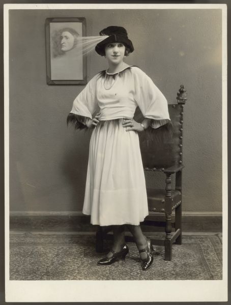 Actress Helen Ferguson poses in a white dress trimmed with monkey fur and black hat with a large feather.  She stands in front of a chair with her hands on her hips.  A framed picture of a man's head is on the wall behind her.