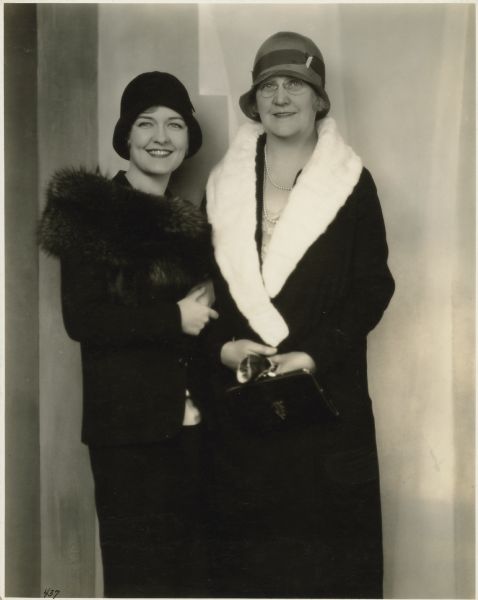 Actress Laura La Plante poses with her mother Lydia.  The women both wear dark fur-trimmed coats and hats; Lydia's coat is lined with white fur and she is holding gloves and purse.