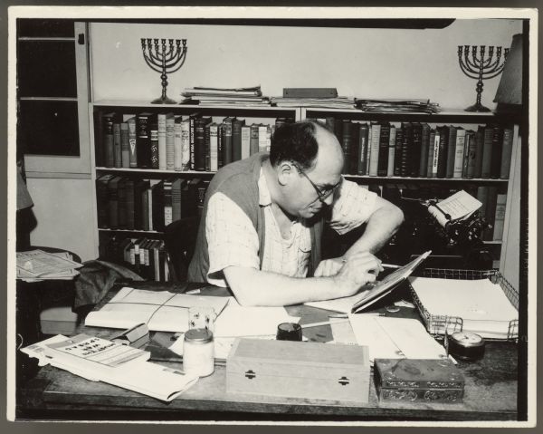 Writer Samuel Ornitz sits at a desk and has one arm resting on a typewriter.  He has a book propped open on the desk which he is looking at.  There are full bookcases behind him with two menorah. The book <i>Must the War Spread</i>, published in January 1, 1940, is on the corner of the desk.