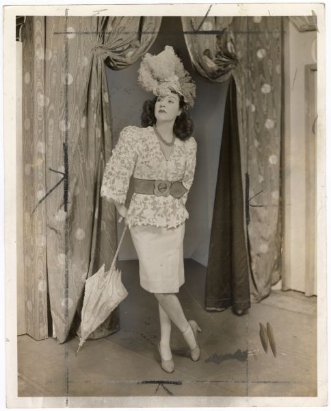 Ethel Merman poses onstage for a publicity photograph for the musical <i>Du Barry Was a Lady</i>.  She wears a blouse and skirt with a large belt and a hat with a large plume while partially leaning on a parasol.  The curtains behind her are pulled open enough for someone to walk through them.