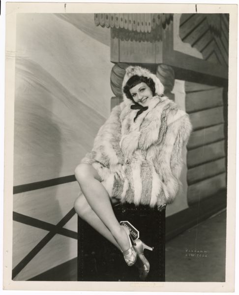 Actress Mary Martin sits cross-legged on top of a trunk in a publicity photograph for the musical <i>Leave It to Me!</i>.  She is wearing a fur coat that covers her body except for her legs. Painted scenery can be seen behind her.