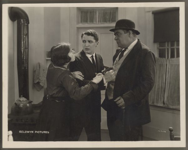 Helen Ferguson grabs onto Bryant Washburn in a scene from the 1922 film <i>Hungry Hearts</i>.  Washburn is next to a much larger man who is wearing a suit and a hat.  Both men look at Ferguson.  The three are standing in a kitchen