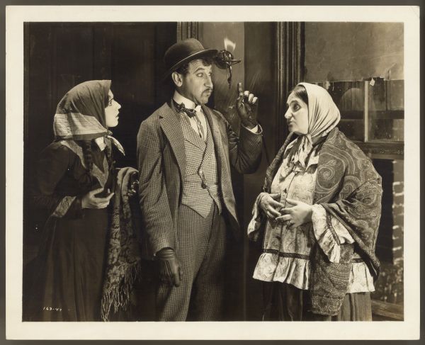 Helen Ferguson, Otto Lederer, and Rosa Rosanova appear in a scene from the 1922 film <i>Hungry Hearts</i>.  Ferguson and Rosanova look at Lederer who is making a face and pointing upwards.  The women both have head scarves on and are either holding or wearing a blanket. Lederer wears a hat and gloves.