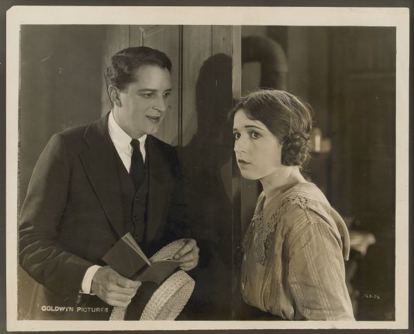 Bryant Washburn stands in a doorway and looks at Helen Ferguson in a scene from the 1922 film <i>Hungry Hearts</i>.  Washburn holds a straw hat and small book and smiles slightly while looking at Ferguson who is looking away from him.