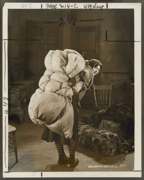 Helen Ferguson carries a large bundle of material on her back in a still for the 1922 film <i>Hungry Hearts</i>.  She has her back to the camera and looks over her shoulder.  There is another large bundle and suitcases behind her in what appears to be a kitchen.