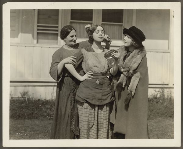 Helen Ferguson holds a mirror and looks at herself while Rosa Rosanova and Ferguson's sister Catherine fix her hair and put makeup on.  Helen and Rosanova are in costumes for the 1922 film <i>Hungry Hearts</i>.  Catherine wears a coat with fur trim.